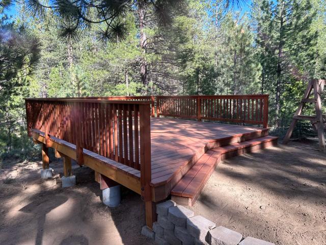 Freestanding redwood deck with picket railing and wraparound steps among tall pines